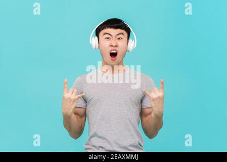 Divertente uomo asiatico che indossa le cuffie e si diverte ad ascoltare le sue canzoni preferite si isolarono su uno sfondo di studio azzurro Foto Stock