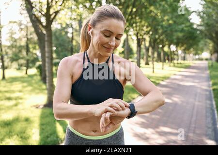 Jog di mattina. Bella donna sportiva in abbigliamento sportivo e auricolari guardando il suo orologio, controllando il risultato mentre corre in un parco verde su un soleggiato Foto Stock