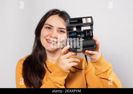 Una bella bruna donna tiene in mano una macchina fotografica Polaroid e sorride su uno sfondo bianco. Formazione fotografica, corsi di fotografo. Foto Stock