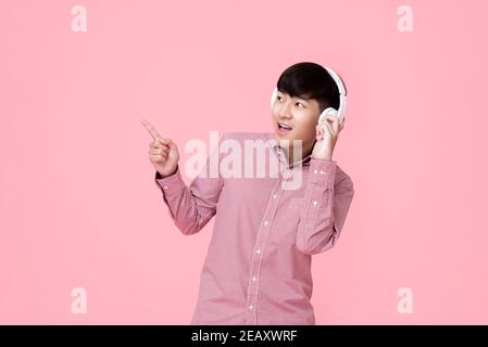 Felice giovane asiatico che indossa cuffie wireless per ascoltare la musica con la mano che punta allo spazio vuoto accanto studio girato isolato su sfondo rosa Foto Stock