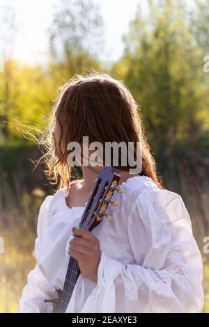 Vista posteriore di giovane donna con capelli rossi in un vestito bianco medievale. Ragazza hippie che tiene la chitarra acustica alla natura. Foto Stock