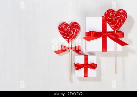 Divertimento valentines giorno sfondo - scatole regalo bianche con arco di seta rossa, cuori di lecca dolce su tavola di legno bianco, spazio copia. Foto Stock