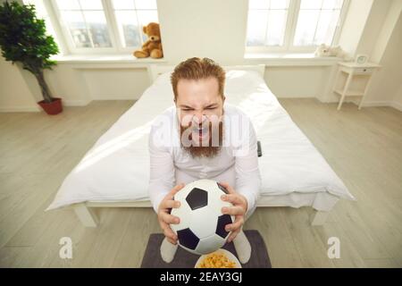Uomo divertente in pigiama bianca seduto con la palla di calcio dentro mani sul letto e gridare Foto Stock