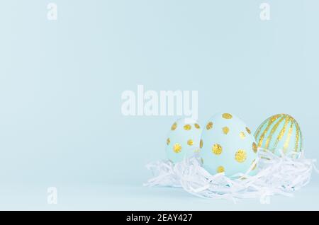 Uova di pasqua blu con puntini di glitter oro in nido bianco su sfondo blu, spazio copia. Foto Stock