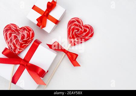 San Valentino sfondo giorno - scatole regalo bianche con nastro di seta rossa e cuori di lecca su tavola di legno bianco come bordo, primo piano. Foto Stock