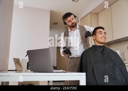 Parrucchiere che fa taglio di capelli con uno strumento speciale per piacere cliente in appartamenti Foto Stock