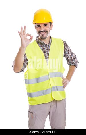 Felice costruttore maschile in elmetto giallo e gilet riflettente in piedi con la mano sulla vita su sfondo bianco e mostrando ok segno mentre si guarda la camma Foto Stock