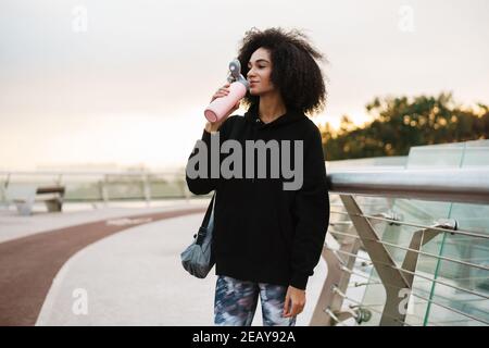 Attraente giovane sportiva africana che trasporta il tappeto di idoneità e tiene l'acqua bottiglia camminare all'aperto Foto Stock