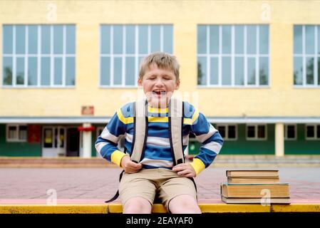 triste scolaro dispiaciuto, il ragazzo grida e non vuole andare a scuola, all'aperto di giorno. Seduto sui gradini con libri e un grande Foto Stock