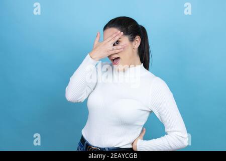 Giovane donna caucasica che indossa un maglione bianco su sfondo blu sbirciando in shock coprendo viso e occhi con la mano, guardando attraverso le dita con embar Foto Stock