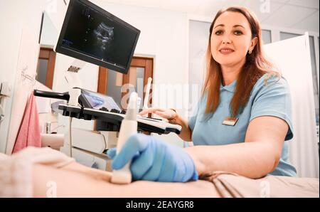 Donna ecografa in guanti sterili che esamina il paziente con scanner a ultrasuoni. Medico che guarda la fotocamera e sorride durante l'esame ecografico. Concetto di sonografia e diagnostica a ultrasuoni Foto Stock