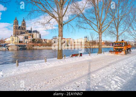 La macchina spazzaneve pulisce una strada dalla neve lungo il fiume Elba e la Cattedrale nel centro storico in inverno a cielo blu e giorno di sole, Magdeburg, Foto Stock