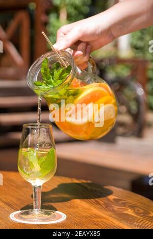 presa a mano femminile e versamento da brocca di vino bianco estivo sangria con frutta a un bicchiere sulla terrazza estiva del ristorante Foto Stock
