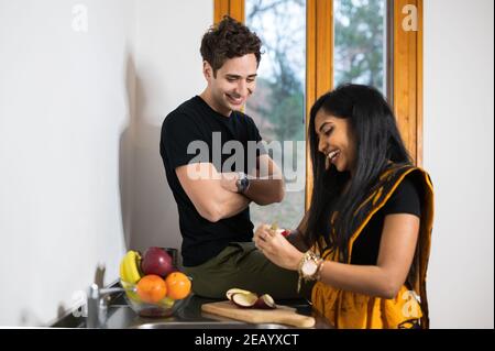 Giovane coppia multietnica a casa. Donna indiana con abito tradizionale che prepara frutta mentre parla con l'uomo caucasico seduto sul banco della cucina Foto Stock