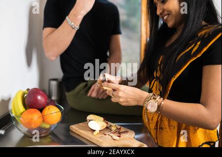 Primo piano donna indiana con abito tradizionale in cucina che spellano una mela, mentre l'uomo caucasico irriconoscibile seduto sul banco. Vita a casa conce Foto Stock