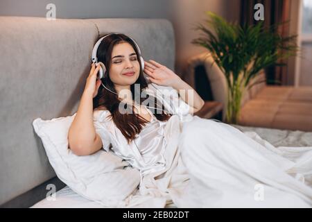 donna ascolta la sua canzone preferita con le cuffie. mattina o sera a letto di una giovane donna con le cuffie Foto Stock