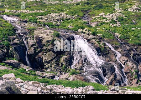 Rapido ruscello in valle di montagna tra le rive erbose. Piccola cascata in prato verde. Foto Stock