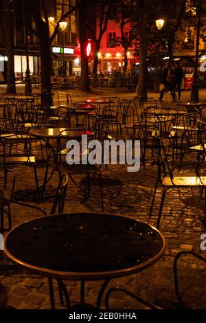 Scena urbana notturna parigina dopo la pioggia. Luci sfocate di bar e negozi che si riflettono sui tavoli vuoti e bagnati da caffè e sul pavimento in ciottoli sulla piazza. Foto Stock