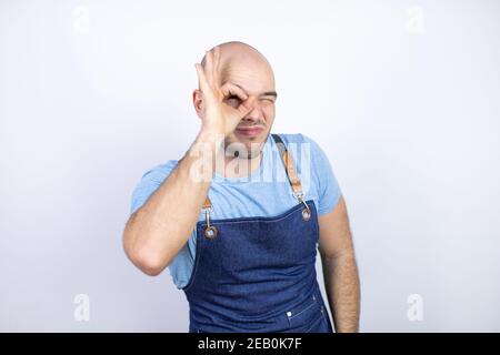 Giovane uomo calvo che indossa grembiule uniforme su isolato sfondo bianco facendo gesto ok scioccato con viso sorridente, occhio guardando attraverso le dita Foto Stock