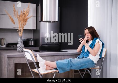 Donna annoiata e sbavata guardando i media sul telefono cellulare seduto su una sedia in cucina a casa Foto Stock