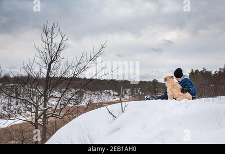 Teen boy e il suo cane seduti sulla cima di una collina innevata il giorno d'inverno. Foto Stock