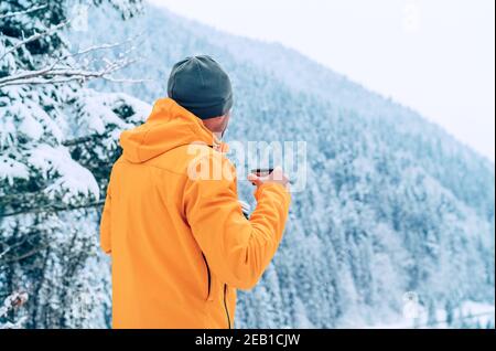 Uomo con tazza bere una bevanda calda vestito giacca softshell arancione brillante e godendo di paesaggi di montagne innevate mentre trekking percorso invernale. Attivo Foto Stock