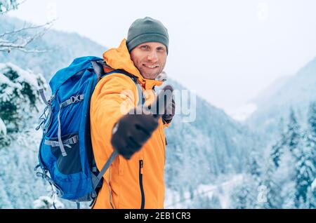 Giacca Softshell Raughing Man vestita arancione brillante con lo zaino alla macchina fotografica e mostrando un gesto di pollice-su mentre lui trekking montagne invernali Foto Stock