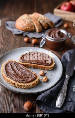 Colazione sana, fette di pane integrale con mela spalmata di cacao in polvere, nocciole tostate macinate e cioccolato fondente su legno rustico Foto Stock
