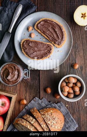 Colazione sana, fette di pane integrale con mela spalmata di cacao in polvere, nocciole tostate macinate e cioccolato fondente su legno rustico Foto Stock