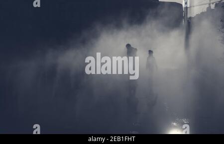 Silhouette sfocate di uomo e ragazza irriconoscibili che giocano nella nebbia d'acqua della fontana il giorno d'autunno. Les Halles Square, Parigi, Francia. Foto scura a toni. Foto Stock