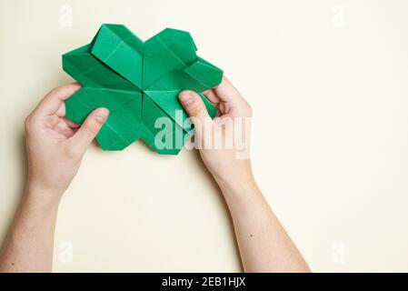 Trifoglio a quattro foglie di carta, origami, tenuto da due mani di giovane donna su uno sfondo chiaro. Concetti di artigianato, fortuna e giorno di San Patrizio. Immagine Foto Stock