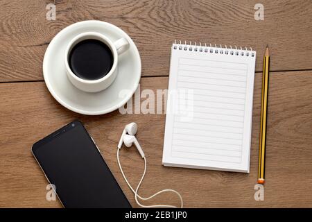 Notebook vuoto accanto a una matita, una tazza di caffè e telefono con cuffie. Tutto su un tavolo di legno. Speciale per mockup. Foto Stock