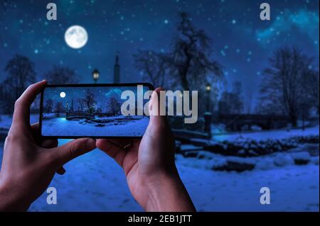 Turista fotografare la magica notte invernale con cielo stellato e luna piena in un parco innevato con un bel ponte sopra un piccolo stagno, luce di strada e coperto Foto Stock