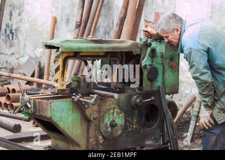 Un lavoratore seghe una billetta d'acciaio su una vecchia macchiata d'olio sega a nastro Foto Stock