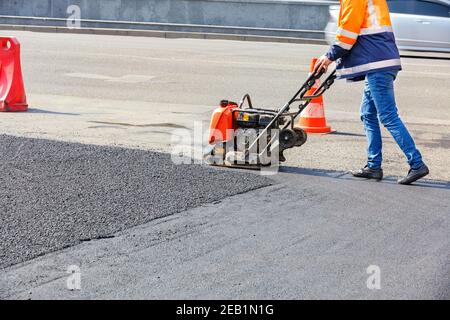 Un operatore su strada utilizza una piastra vibrante per compattare l'asfalto su un sito di riparazione stradale recintato in una giornata di sole. Foto Stock