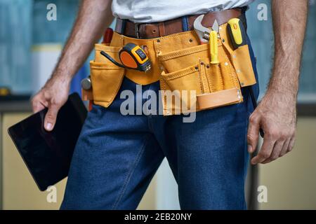 Un colpo corto di giovane riparatore che indossa una cintura portautensili con vari strumenti che tengono il tablet digitale in piedi in interni Foto Stock