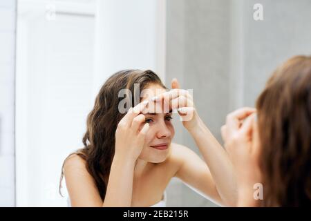 Giovane donna che guarda e stringe l'acne su una faccia davanti allo specchio. Brutto problema di pelle ragazza, ragazza teen che ha pimples. Cura della pelle. Bellezza Foto Stock