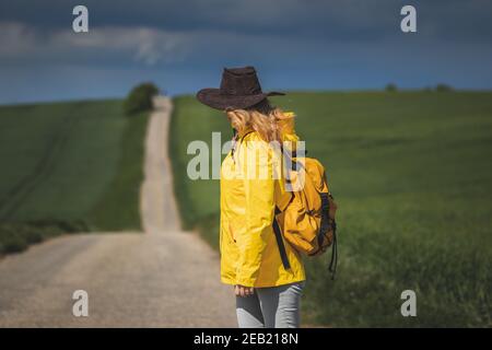 Donna che camminano sulla strada e guardando il cielo nuvoloso. La pioggia sta arrivando. Zaino in spalla con cappello e giacca impermeabile gialla Foto Stock