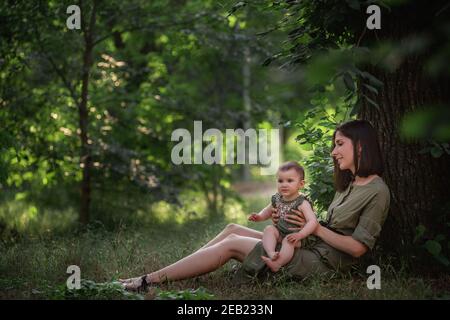 Una giovane madre sana tiene un bambino piccolo nelle sue braccia. Una famiglia felice siede sull'erba verde, sotto un albero alto, gioca, abbracci, gode di una passeggiata nel Foto Stock