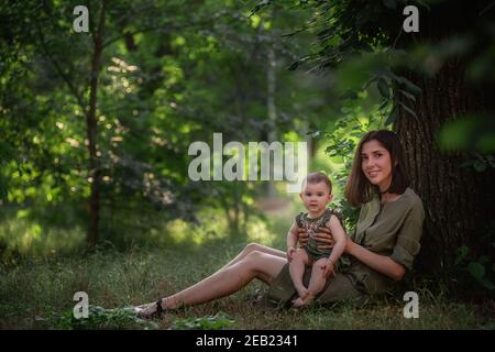 Una giovane madre sana tiene un bambino piccolo nelle sue braccia. Una famiglia felice siede sull'erba verde, sotto un albero alto, gioca, abbracci, gode di una passeggiata nel Foto Stock