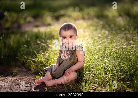 Un piccolo bambino carino con i capelli corti, in un vestito verde si siede sul prato in erba vicino al sentiero, guarda in su nel cielo al sole. Lei sorride a lei Foto Stock