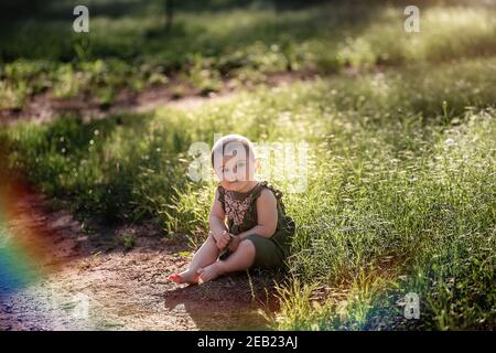 Un piccolo bambino carino con i capelli corti, in un vestito verde si siede sul prato in erba vicino al sentiero, guarda in su nel cielo al sole. Lei sorride a lei Foto Stock