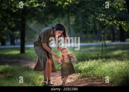 La bambina prende i primi passi, una giovane madre che tiene un bambino per le mani, assicurandosi, aiutando a camminare lungo il percorso del parco in mezzo all'albero verde Foto Stock