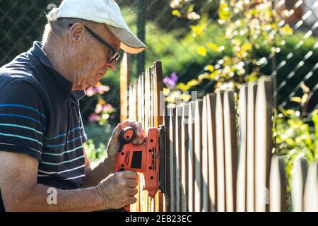 Uomo anziano levigare recinzione in legno con levigatrice elettrica in giardino. Riparazione picket recinto in cortile Foto Stock