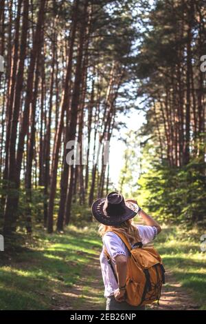Donna con cappello e zaino escursioni in foresta. Escursionista in piedi sul sentiero e guardando nel bosco. Il turista femminile è trekking da solo Foto Stock