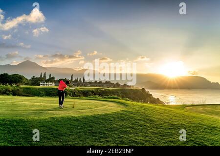 Lone, golfista alla moda, si tee al tramonto su un campo oceanico nei tropici delle Hawaii. Azione divot girato con raggi di sole dorati che si riflettono sull'oceano Foto Stock