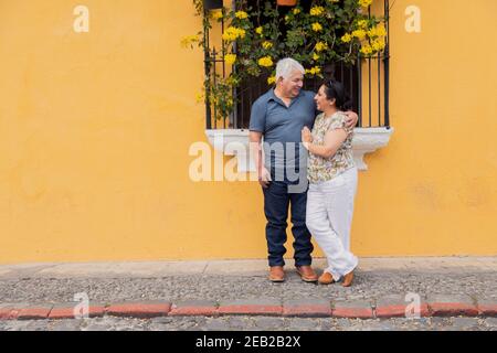 La coppia adulta si abbraccia guardando l'un l'altro su un giallo Muro con fiori in Antigua Guatemala - Coppia senior innamorata in vacanza nella città coloniale Foto Stock