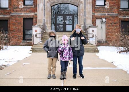 Tre bambini piccoli sono seduti sui gradini anteriori della loro scuola elementare edificio, indossando backpact e mascherine protettive durante il Pandemic. Foto Stock