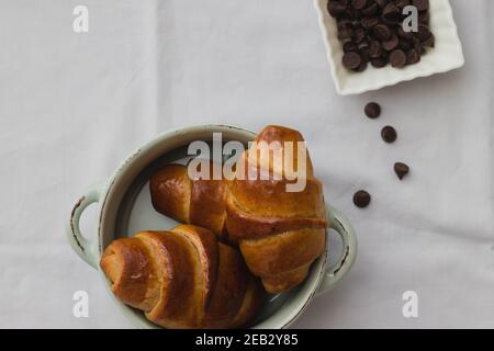 croissant fatti in casa a base di farina intera, in una piccola pentola di ceramica accanto a gocce di cioccolato, su fondo bianco Foto Stock