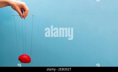 Un cervello di rosso su sfondo blu, una mano che manipola la mente come una marionetta. Il controllo del cervello del concetto. Foto Stock